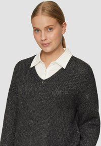 Dunkelgrauer, lockerer Strickpullover mit V-Ausschnitt, strukturiertem Stoff und Metallfäden; getragen über einem weißen Hemd mit Kragen.