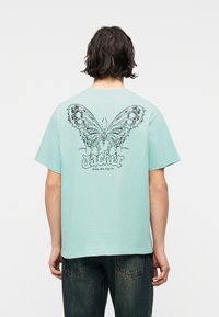 Άτομο με κοντά σκούρα μαλλιά που φοράει ανοιχτό μπλε μπλουζάκι με μεγάλο, λεπτομερές σχέδιο πεταλούδας και κείμενο στην πλάτη, συνδυασμένο με σκούρο τζιν.