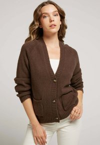 Cardigan en tricot marron avec un col en V, deux poches avant et trois boutons. Texture douce, poignets et ourlet côtelés. Coupe ajustée.