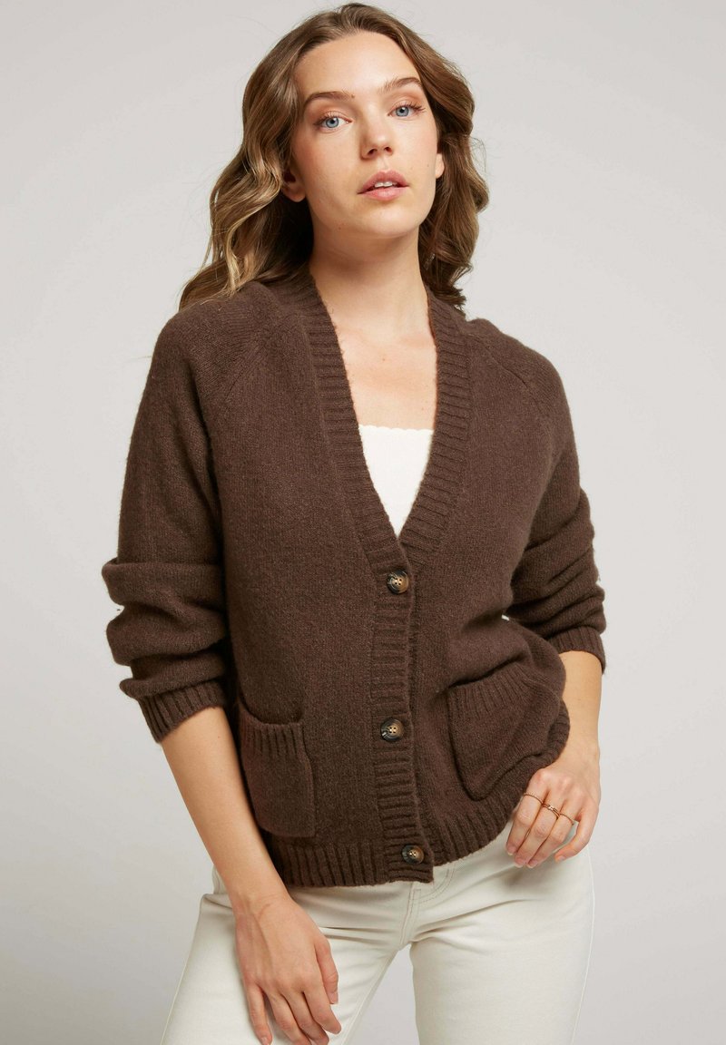Cardigan en tricot marron avec un col en V, deux poches avant et trois boutons. Texture douce, poignets et ourlet côtelés. Coupe ajustée.