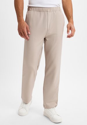 Beige Hose mit elastischem Bund, geradem Schnitt und seitlichen Taschen, kombiniert mit weißen Sneakern auf einem einfarbigen Hintergrund.