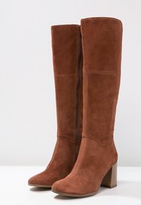 Paire de bottes hautes en daim marron avec fermeture éclair latérale et talons carrés sur une surface blanche contre un fond gris clair.