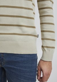 Beige gestreepte trui met bruine horizontale strepen, geribde manchetten en zoom, gedragen over spijkerbroeken. Opvallende zijlabeldetail.