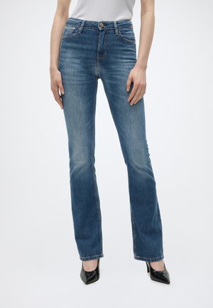 Γυναίκα με μπλε τζιν bootcut, λευκό τοπ και μαύρα μυτερά ψηλοτάκουνα παπούτσια, στέκεται μπροστά σε μία απλή φόντο.
