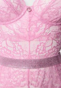 Ροζ δαντελένιο ένδυμα με λουλουδάτα σχέδια, στολισμένο με ένα μικρό κόσμημα σε σχήμα καρδιάς στο κέντρο και μια φαρδιά ζώνη διακοσμημένη με στρας στη μέση.