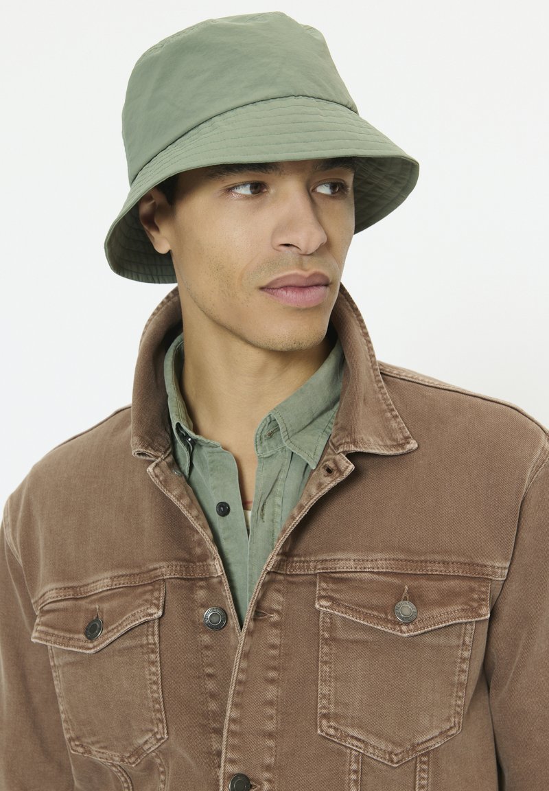 Chapeau bob vert clair avec une finition texturée ; porté avec une veste en denim marron comportant deux poches poitrine et une chemise vert clair en dessous.