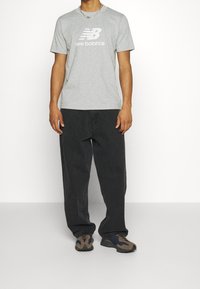 Světlounce šedé tričko s krátkým rukávem a logem "New Balance", doplněné volnými černými džínami a hnědými sportovními botami s texturovaným designem.