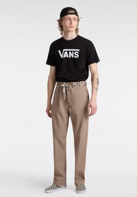 Maglietta nera Vans con grande logo bianco, pantaloni beige con vita elastica e coulisse a fantasia, abbinati a scarpe a scacchi bianche e nere.