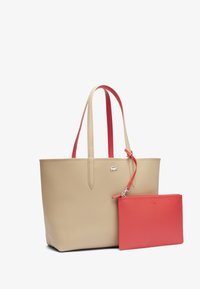 Sac fourre-tout en cuir beige avec des poignées rouges, comprenant une petite pochette en cuir rouge attachée. Le sac a une texture lisse et un détail de logo.
