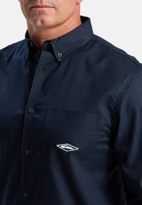 Marineblauw overhemdd shirt gemaakt van katoen, met een borstzak en een geborduurd logo op de zak. Klassieke kraag en gestructureerde stof.