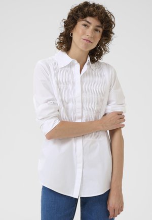 Chemise blanche à manches longues et à boutons, avec une texture plissée sur le devant, des poignets retroussés et un col classique. Associée à un jean bleu.
