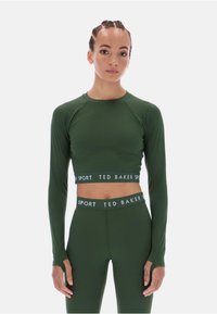 Tamsiai žalia sportinė trumpa palaidinė su ilgomis rankovėmis, pasižyminti balta prekės ženklo elastinga juosta. Lygi, tampri medžiaga su subtiliais tekstūros akcentais.