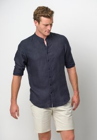 Camicia in lino blu navy con colletto rialzato e maniche a metà. Presenta una chiusura frontale con bottoni ed è abbinata a pantaloni corti beige chiaro.