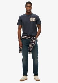 Marinblå bomulls-T-shirt med "Superdry Athletics"-logotyp, ihop med blå denimjeans och en rutmönstrad flanellskjorta knuten i midjan.