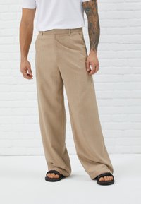 Pantalons beige à jambes larges en tissu texturé, dotés de poches avant et d'une coupe droite, portés avec des tongs noires.