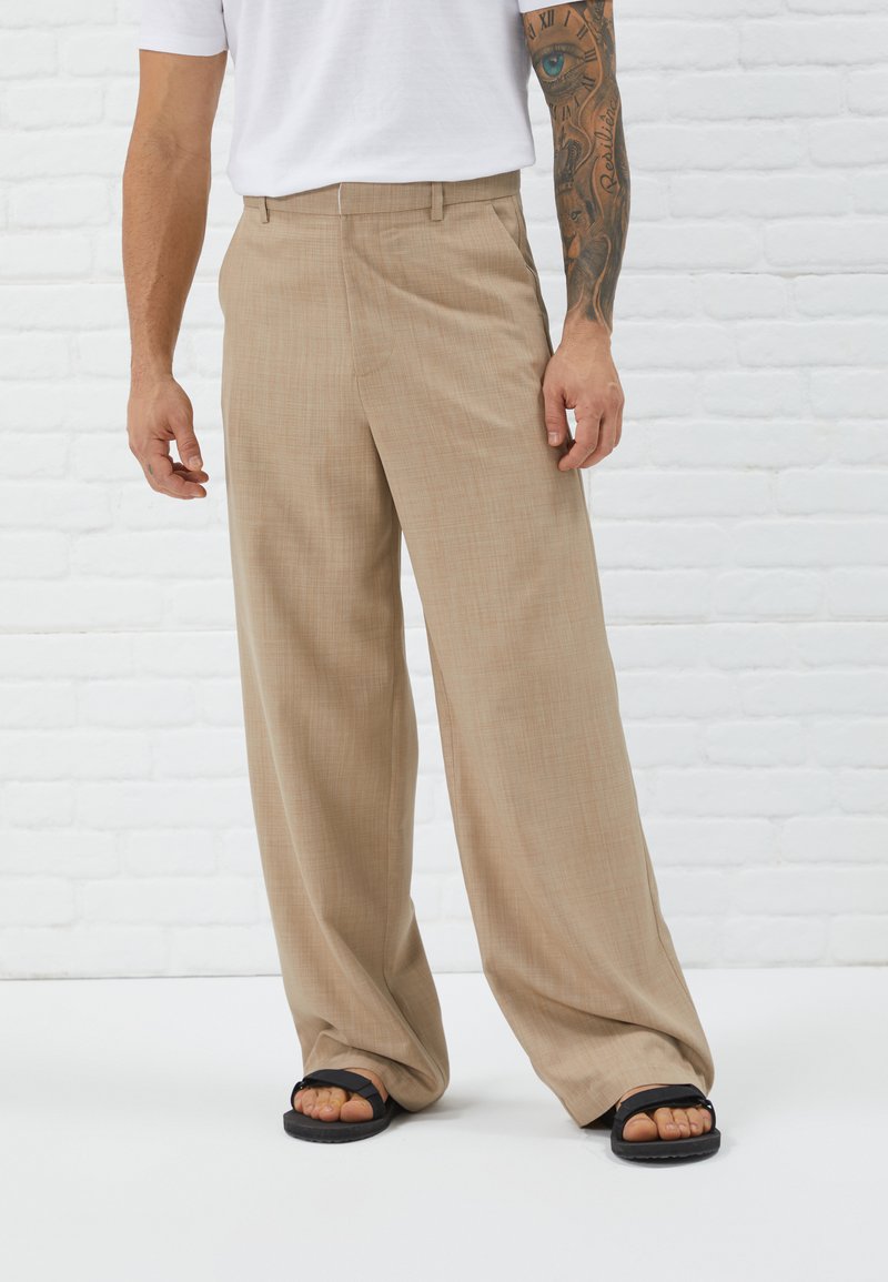 Pantalons beige à jambes larges en tissu texturé, dotés de poches avant et d'une coupe droite, portés avec des tongs noires.