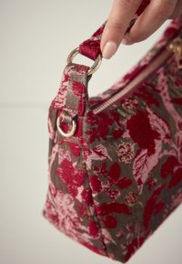 Hand hält eine Stofftasche mit Blumenmuster, roter und rosa Stickerei und goldenem Reißverschluss vor einem einfarbigen Hintergrund.