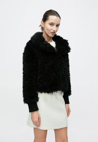 Μαύρο μπουφάν από ψεύτικη γούνα με φαρδύ γιακά, υπερμεγέθη εφαρμογή και ριμπ μανσέτες, σε συνδυασμό με λευκή μίνι φούστα με υφή.