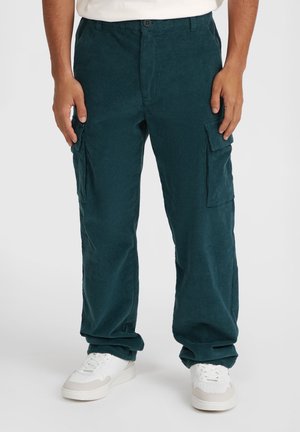 Cargo bukser i mørk teal kordfløjl med to sidelommer; løs pasform med tekstureret overflade, båret med hvide sneakers.