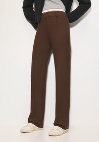 Bruine, op maat gemaakte broeken met een recht model; gladde stof, plooien aan de voorkant en zijzakken; gecombineerd met beige sportschoenen.