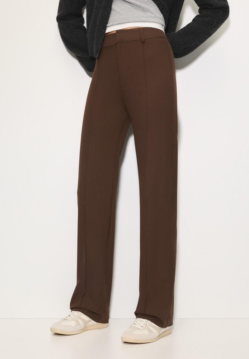 Bruine, op maat gemaakte broeken met een recht model; gladde stof, plooien aan de voorkant en zijzakken; gecombineerd met beige sportschoenen.