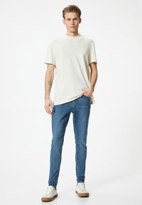 T-shirt a girocollo color beige chiaro abbinata a jeans blu aderenti e sneakers bianche con suole marroni, con una silhouette casual e slim-fit.