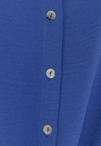 Blue fabric shirt featuring a row of silver buttons, smooth texture, and clean stitching along the button placket.