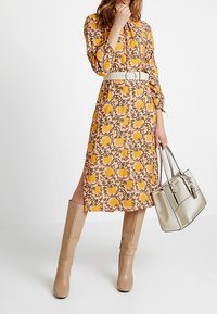 Persikkavärinen midimekko, jossa keltakukkainen kukkakuosi ja vyötärölle istuva valkoinen vyö. Yhdistetty beigeihin polvipitkiin saappaisiin ja hopeanväriseen käsilaukkuun.