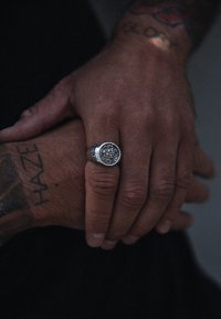 Sidabrinis žiedas su išsamiu dizainu, kuriame yra geometrinių raštų, uždėtas ant tatuiruotą ranką, kuri ilsisi ant kitos rankos.