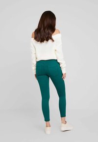 Pantalones vaqueros ajustados verdes con un diseño entallado, que cuentan con bolsillos traseros. Combinados con un suéter de punto blanco suelto y con hombros al descubiertos, y zapatillas de color claro.