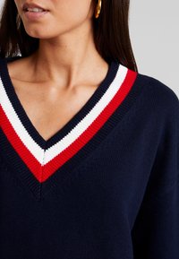 Marineblaues Sweater mit V-Ausschnitt, der einen gerippten Kragen in Rot, Weiß und roten Streifen aufweist. Stricktextur, körperbetonter Schnitt.