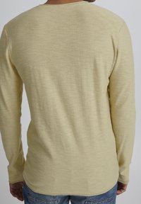 Vue arrière d'une personne portant une chemise à manches longues jaune clair et un jean bleu, sur fond uni.