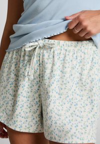 Woman wearing white shorts with small blue floral print and a light blue ribbed top, holding the hem to reveal waist area.