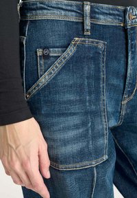 Jeans de mezclilla azul oscuro con un ajuste slim, costuras visibles y un bolsillo frontal. El material parece duradero con un ligero desgaste.