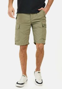 Cargo shorts i olivengrøn. Letvægtsstof, knælang, flere lommer og et logo-patch på venstre side. Cremerfarvede sneakers bæres.
