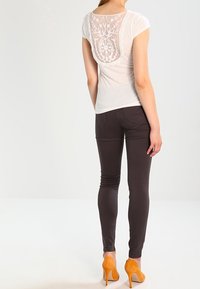 Bílá tričko s krajkovým detailem na zádech, kombinované s tmavě hnědými slim-fit kalhotami a oranžovými vysokými podpatky. Hladká textura a přiléhavý design.