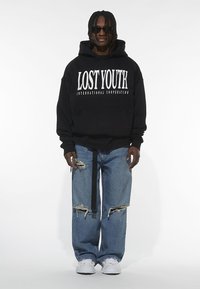 Black hooded sweatshirt with white text "LOST YOUTH INTERNATIONAL COOPERATION," paired with ripped blue jeans and white sneakers. Black sunglasses.
