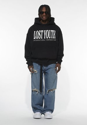Black hooded sweatshirt with white text "LOST YOUTH INTERNATIONAL COOPERATION," paired with ripped blue jeans and white sneakers. Black sunglasses.