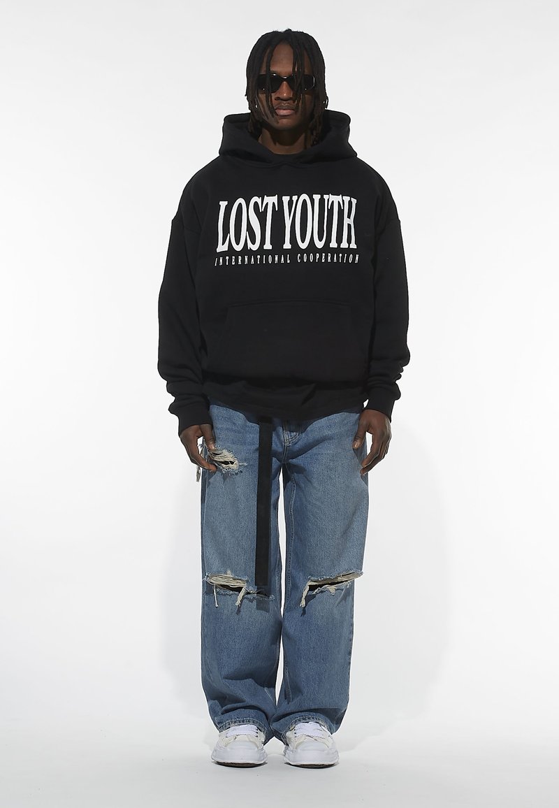 Black hooded sweatshirt with white text "LOST YOUTH INTERNATIONAL COOPERATION," paired with ripped blue jeans and white sneakers. Black sunglasses.