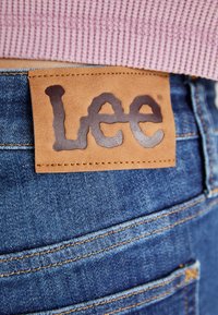 Étiquette en cuir marron avec le logo "Lee" cousue sur un jean en denim bleu porté avec un pull en maille rose visible à la taille.