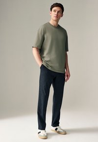 Olivengrøn oversized t-shirt med rund hals, kombineret med mørkegrå teksturerede bukser og hvide sneakers med sorte detaljer.