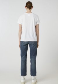 Bílé bavlněné tričko s krátkými rukávy, doplněné modrými džíny z denimu. Model má na sobě bílé tenisky a je zobrazen ze zadní strany.