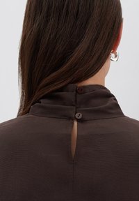 Donna con capelli brown lisci, che indossa orecchini a cerchio argentati e un top marrone scuro abbottonato sulla nuca.
