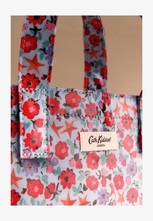 Prim-plan cu o geantă din material imprimat floral cu flori roșii, roz și mov și forme stelate, având o etichetă cusută "Cath Kidston London".
