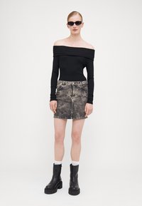 Svart stripete off-shoulder topp, høyt liv, falmet denimskjørt, kraftige svarte støvler, og mørke solbriller, satt mot en nøytral bakgrunn.