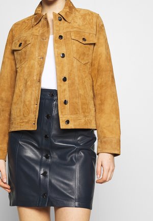 Femme portant une veste en daim beige avec des boutons noirs sur un haut blanc, associée à une jupe en cuir noire taille haute à boutons.