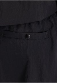 Gros plan d’un pantalon en tissu noir avec une seule poche arrière sécurisée par un bouton noir gravé de "ALPHA".