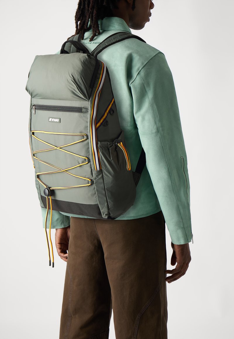 Sac à dos en nylon vert au design structuré, avec des accents contrastants jaunes et noirs, un détail en cordon élastique et une poche zippée à l'avant.