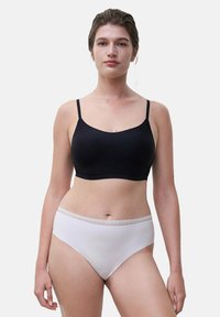 Woman wearing a black spaghetti strap bra and white high-cut lace-trimmed underwear, standing against a plain light background.