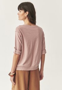 Maglione lavorato a maglia rosa chiaro con maniche a tre quarti, polsini arrotolati fissati da un bottone e una vestibilità morbida. Indossato con pantaloni marrone chiaro.